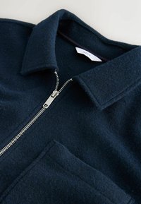 Námornícke modrá bunda z vlnenej zmesi s golierom, predným zipsom a jednou kapsou. Textúrovaná látka a strieborné zipsové kovanie.