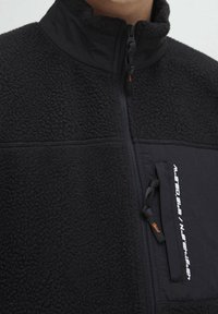 Černá fleecová bunda s vysokým límcem, předním zipem a zipovou kapsou na hrudi s bílým textem a malou oranžovou visačkou.