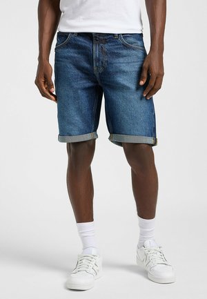 Denim shorts in dark blue, featuring rolled cuffs, five pockets, and a sturdy waistband. Paired with white sneakers and socks.