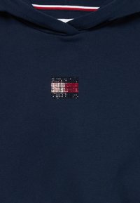 Námornícky modrá mikina s kapucňou a malým logom zdobeným kamienkami. Má šnurovanie na kapucni a textúrovanú látku. Jednoduchý, neformálny dizajn.