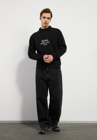 Svart genser med krage, med hvit brodert tekst. Kombinert med løstsittende mørke jeans og svarte sneakers.