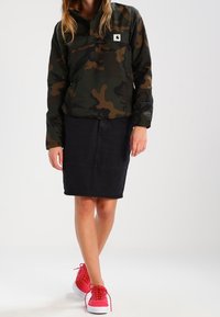 Jakke med camouflage-mønster i mørkegrøn og brun, sort denimnederdel og lyserøde sneakers med hvide såler og snørebånd.