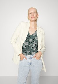 Blazer ajustado off-white sobre um top verde escuro com padrões florais brancos, combinado com jeans de ganga azuis claros.