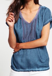 Niebieska bluzka z krótkim rękawem i dekoltem w serek, z misternymi pionowymi przeszyciami oraz kontrastującymi fioletowymi akcentami wzdłuż dekoltu. Miękka faktura materiału.