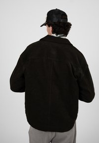 Mann trägt eine schwarze Cordjacke, graue Hose, gemusterten Schal und schwarze Netz-Baseballkappe, blickt mit dem Rücken zum Betrachter vor einem einfarbigen grauen Hintergrund.