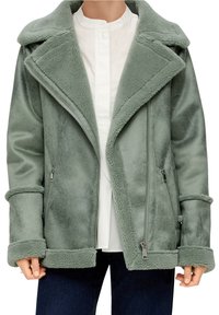 Persoon met een lichtgroene schapenvachten jack met een oversized kraag, ritszakken en opgerolde mouwen, gedragen over een wit shirt en donkere broek.