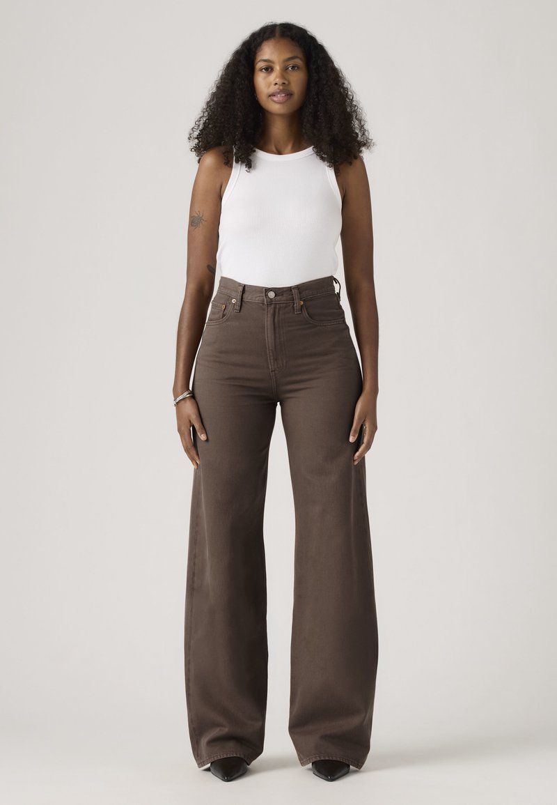 Jeune femme aux cheveux bouclés portant un haut blanc sans manches et un pantalon large taille haute marron, debout avec les bras détendus le long du corps.