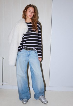 Young woman with long hair wears striped long-sleeve shirt, light wide jeans, silver sneakers, and white fluffy coat draped over one shoulder.