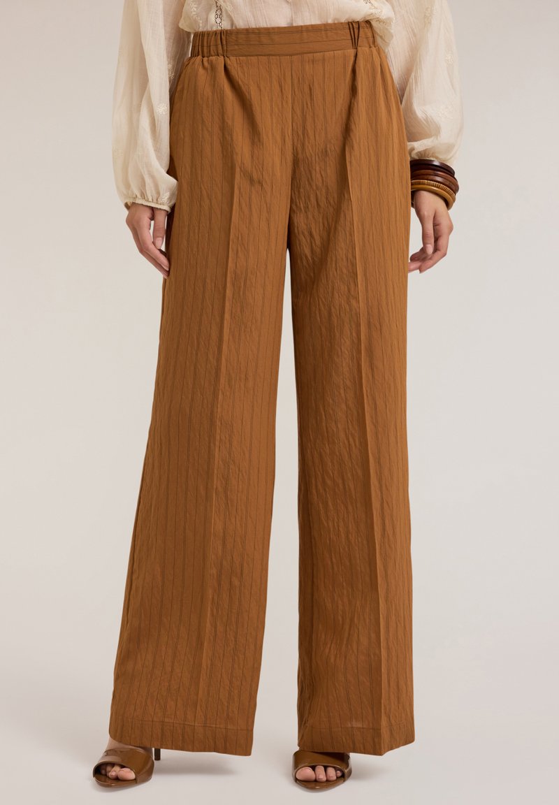 Brede broek in een warme bruine kleur, met verticale streepjes en een textuurstof. Elastische tailleband, gecombineerd met open neus hakken.