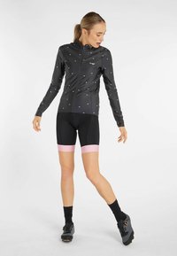 Black, slim-fit cycling jacket with silver eye patterns, full zip, high collar, and long sleeves. Paired with black shorts and pink accents.