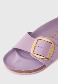 Vaaleanlila Birkenstock-sandaali, jossa on leveä hihna, kultainen solki ja korkkipohja. Sujuva pinta, muotoiltu pohja ja säädettävä suljenta.
