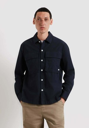 Giovane uomo con capelli corti neri e lentiggini che indossa una camicia nera abbottonata con tasche sul petto e pantaloni beige, in piedi con le mani in tasca.