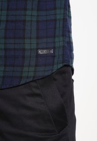 Geruite blouse in donkerblauw en groen flanel met een gebogen zoom, voorzien van een klein zwart label aan de onderkant. Gecombineerd met een zwarte broek.
