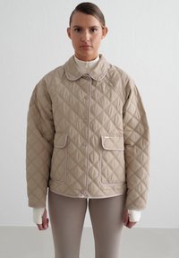 Beige quiltet jakke med rund krave, trykknapper, to frontlommer og kontrasterende lys trim. Brukt over en tettsittende topp og leggings.