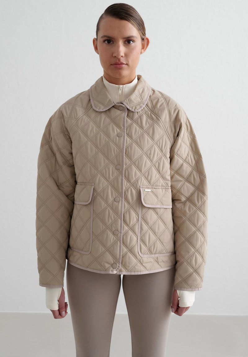 Beige quiltet jakke med rund krave, trykknapper, to frontlommer og kontrasterende lys trim. Brukt over en tettsittende topp og leggings.