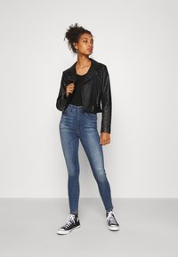 Veste de moto en cuir noir, top noir côtelé, jeans skinny taille haute bleu, et baskets en toile noir et blanc avec des accents rayés.