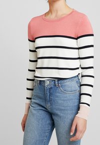 Kvinna klädd i en långärmad tröja med rund halsringning, rosa topp, vit kropp med svarta horisontella ränder, och ljusblå högmidjade jeans.