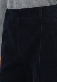 Sötétkék bordázott kordnadrág, lapos elülső résszel és övhurkokkal, világosszürke felső ellenében bemutatva.