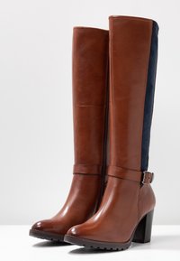 Las botas altas de cuero marrón cuentan con un acabado suave, punta redondeada y tacón cuadrado. Tienen cremallera lateral y detalles en ante azul marino.