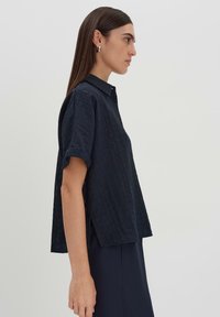 Camicia a maniche corte blu navy con bottoni, realizzata in un tessuto testurizzato e dotata di colletto. Presenta una vestibilità ampia e dettagli con spacco laterale.