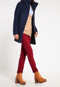 Cappotto di lana blu navy con cappuccio, cardigan beige, pantaloni rossi e stivaletti con tacco marroni. Bottoni visibili e dettagli di tessuto texturizzato.