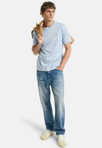 Lichtblauwe katoenen t-shirt met een ronde hals, gecombineerd met vervaagde blauwe jeans en crèmekleurige sneakers. Een tan kleurige jas over één arm gedrapeerd.