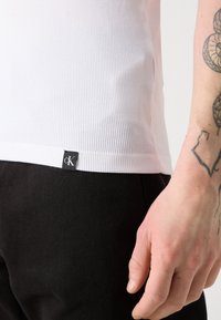 Bijela rebrasta majica bez rukava s crnom "ck" oznakom na rubu, u kombinaciji s crnim hlačama. Vidljiva koža i tetovirana ruka.