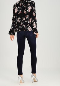 Blouse fleurie noire à manches évasées, associée à un jean skinny bleu foncé et des chaussures à talons hauts argentées. Le haut présente un détail de couture au dos.