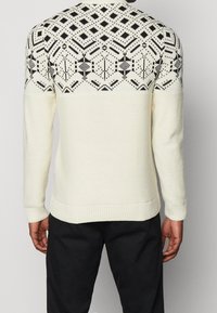 Crème trui met een zwart geometrisch patroon op de bovenrug. Gebreide stof, geribde cuffs en zoom, ontspannen pasvorm. Textuurdetail in het ontwerp.