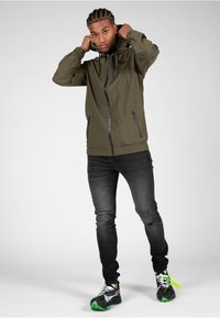 Olivenfarbene Kapuzenjacke mit Reißverschlusstaschen, kombiniert mit schwarzen Skinny-Jeans. Die Schuhe weisen Design-Elemente in Schwarz, Weiß und Grün auf.