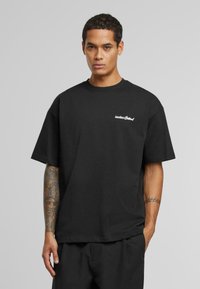 Sort bomulds t-shirt med en afslappet pasform, korte ærmer og et hvidt "Another" logo på venstre bryst. Minimalistisk design, glat tekstur.