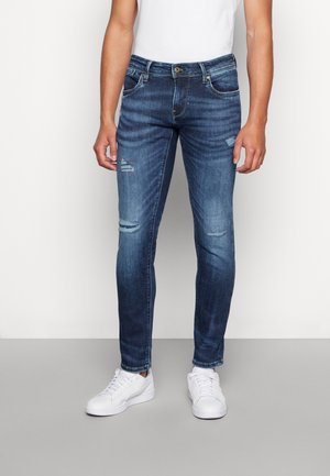 Homme portant un jean bleu délavé slim avec de petites déchirures aux cuisses et aux genoux, des baskets blanches et un t-shirt blanc sur un fond uni.