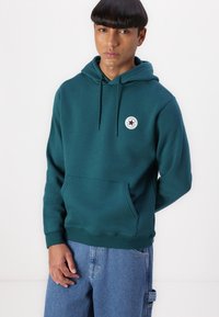 Teal hettegenser i mykt stoff med kengurulomme og en sirkulær logo detalj på brystet, kombinert med løstsittende jeans.
