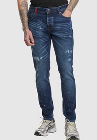 Jeans de denim en un lavado oscuro con desgastes en azul claro, diseño ajustado, cinco bolsillos y un acento rojo en la cinturilla.