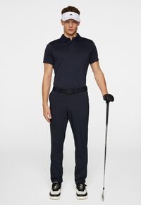 Marinblå polo-shirt och byxor med ett svart bälte, i kombination med svart och vita sneakers. Personen håller en golfklubba och bär en vit skugghatt.