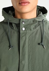 Veste à capuche verte en tissu texturé, dotée de boutons-pression, d'un cordon de serrage ajustable et d'une coupe décontractée autour du cou.