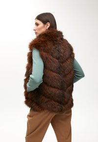 Hnědá kožichová vesta s polstrovanými detaily; textura vypadá měkce a nadýchaně. Nositá přes světle modrý top s dlouhým rukávem a hnědé kalhoty.