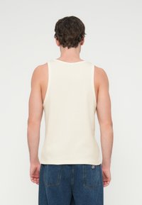 Εκρού αμάνικη μπλούζα με ριμπ υφή, φαρδιές τιράντες και στρογγυλή λαιμόκοψη, σε συνδυασμό με μπλε τζιν. Εμφανίζεται η πίσω όψη.