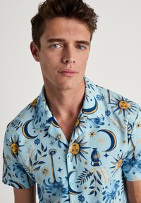 Kortlopende overhemd met kraag in lichtblauwe stof, voorzien van zon-, maan- en sterrenpatronen in geel en marineblauw. Gladde textuur, met knoopsluiting aan de voorkant.