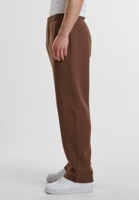 Brune svedpants med elastisk talje set i profil, med en afslappet pasform og glat tekstur, parret med hvide sneakers.