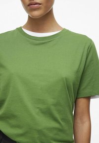Camiseta de algodón verde de manga corta con cuello redondo. Presenta un interior de cuello blanco en contraste y un corte holgado. Textura suave.