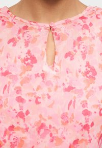 Κοντινό πλάνο ροζ φλοράλ μπλούζας με ένα κουμπί και άνοιγμα σε σχήμα κλειδιού στο λαιμό.