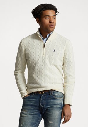 Witte kabelgebreide pullovertrui met een kwart-rits kraag, voorzien van een marineblauw logo op de borst, gecombineerd met versleten blauwe jeans.