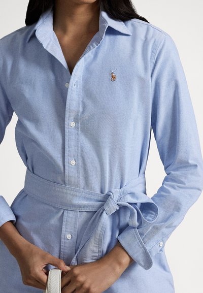 Kvinne iført en lys blå skjortekjole med knapper og belte i livet, som holder en hvit stropp i hendene.