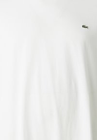 Vit V-ringad skjorta med en liten grön krokodil logotyp broderad på vänster bröst.