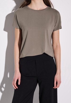 Kobieta ubrana w gładki taupe krótki rękaw t-shirt, lekko włożoną w czarne spodnie z przednimi kieszeniami, na tle białej ściany.