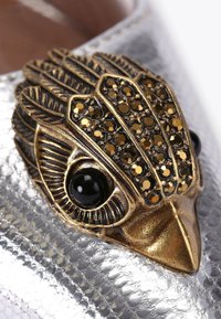 Chaussure en argent métallique ornée d'un accent en bronze en forme d'oiseau, présentant des plumes texturées et des gemstones noirs pour les yeux.