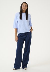 Pull en maille bleu clair avec un motif de feuilles texturé et une coupe décontractée, associé à un jean large bleu foncé et des chaussures de couleur claire.