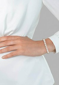Weißes Perlenarmband an einem Handgelenk, das gleichmäßig angeordnete Perlen und eine glatte Textur aufweist, getragen vor einem hellen Stoffhintergrund.