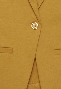 Gros plan sur un blazer jaune moutarde avec un seul bouton beige fermé, un col châle et deux poches passepoilées horizontales.